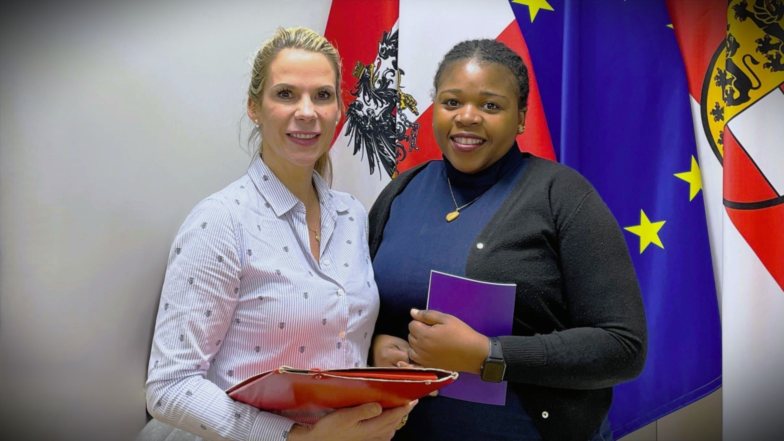 Zwei Frauen stehen vor &ouml;sterreichischen und EU-Flaggen und 
halten Unterlagen in den H&auml;nden. Die Szene vermittelt den 
Kontext eines Austauschs und symbolisiert Weiterbildung 
sowie lebenslanges Lernen.