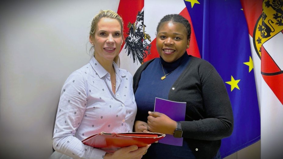 Zwei Frauen stehen vor &ouml;sterreichischen und EU-Flaggen und 
halten Unterlagen in den H&auml;nden. Die Szene vermittelt den 
Kontext eines Austauschs und symbolisiert Weiterbildung 
sowie lebenslanges Lernen.