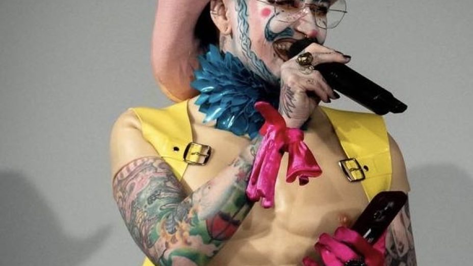 Das Bild zeigt Eric BigClit. Er trägt einen rosa Cowboyhut und hat ein aufwendiges Make-up mit blauen und weißen Akzenten im Gesicht. Er trägt über dem nackten Oberkörper ein gelbes Gilet und an der linken Hand einen pinken 